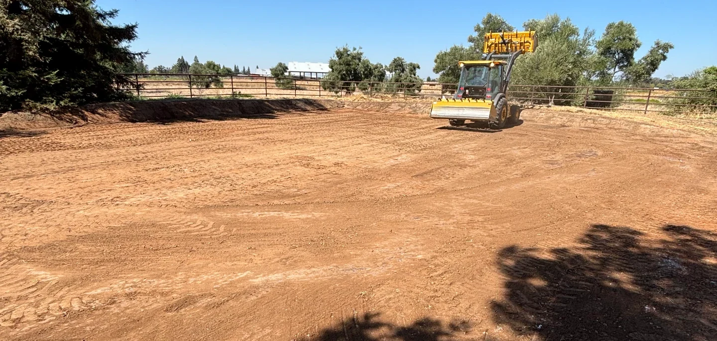Tractor with an arena groomer working a graded dirt arena beside a pipe fence